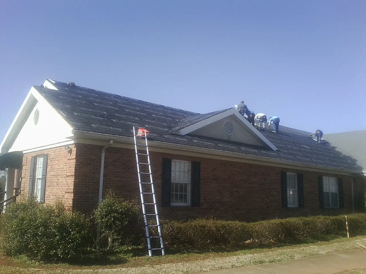 Crew tearing shingles off a brick ranch roof with a ladder set — Emergency Roof Repair work by our crew in North Liberty
